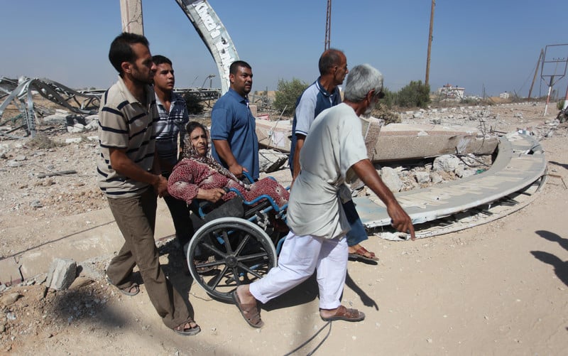 a group of men assisting an elderly woman in a wheelchair through an area with significant destruction and rubble (gaza). The background shows damaged structures and debris, suggesting the scene is in a post-conflict or disaster area. The men appear focused and determined to help the woman navigate the challenging terrain.