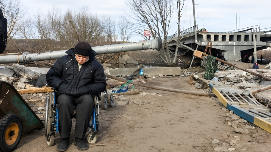 an elderly man in a wheelchair in what appears to be a desolate, outdoor setting with debris and fallen structures around him. He is dressed warmly in a winter jacket and a hat, looking downwards, which may suggest a somber or reflective mood. The background indicates a possibly damaged or neglected area, adding to the overall sense of hardship or struggle.