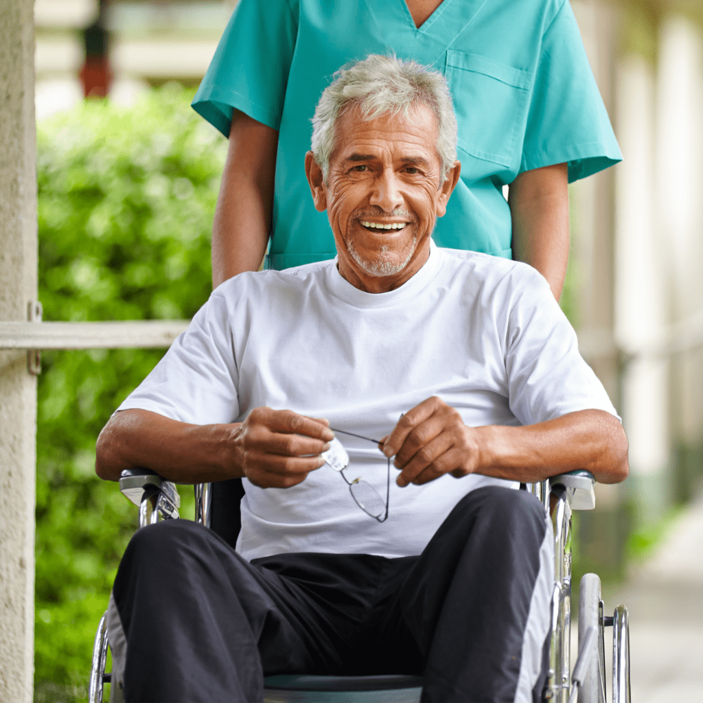 an elderly man in a wheelchair, smiling and holding his glasses. There is a person standing behind him, wearing a teal medical uniform, suggesting they might be a caregiver or healthcare professional. The setting appears to be outdoors, possibly at a healthcare facility or a park.
