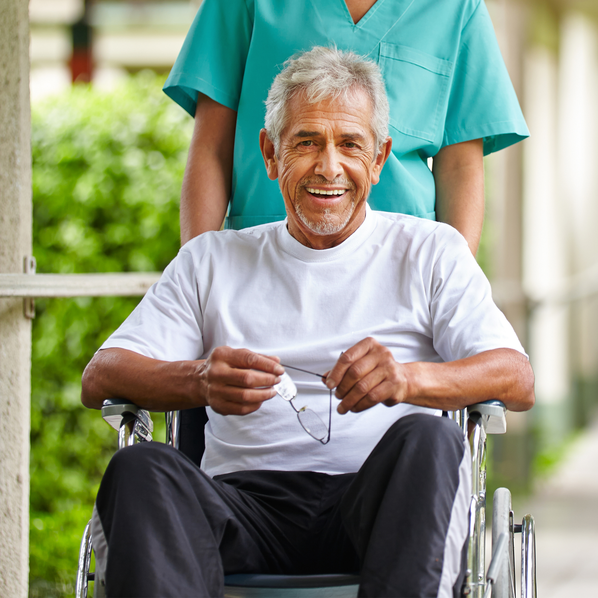 an elderly man in a wheelchair, smiling and holding his glasses. There is a person standing behind him, wearing a teal medical uniform, suggesting they might be a caregiver or healthcare professional. The setting appears to be outdoors, possibly at a healthcare facility or a park.
