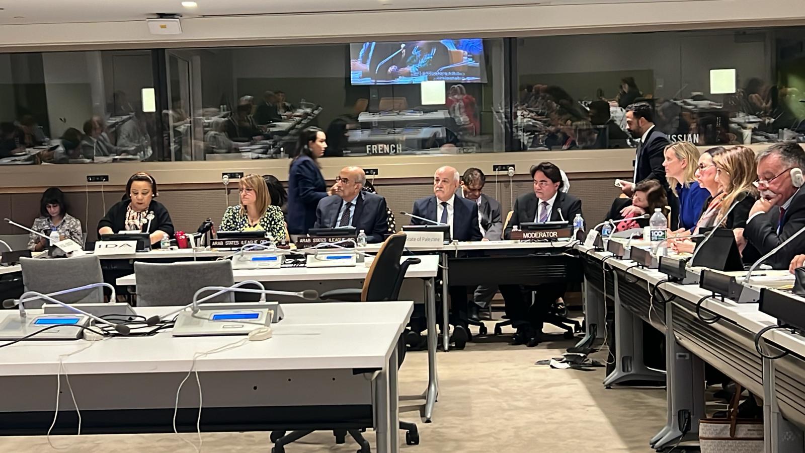 A diverse group of individuals is seated around a large conference table in a formal meeting room. Each person has a microphone and some have nameplates indicating their affiliations, such as "IDA," "Arab States League," and "State of Palestine." The atmosphere suggests a high-level discussion or diplomatic meeting, with various languages being accommodated, as indicated by the labeled glass partition in the background.
