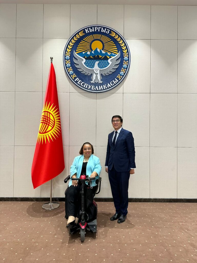 This image shows Dr. Heba Hagras and the Deputy Prime Minister of Kyrgyzstan standing in front of the official government seal and flag, in the presidential building of Kyrgyzstan. The seal on the wall is the national emblem of Kyrgyzstan. It features a golden eagle with spread wings, mountains, a sun, and text in the Kyrgyz language. To the left of the seal is the national flag of Kyrgyzstan, which is red with a yellow sun design in the center. In the foreground are two people: 1. Dr. Heba Hagras in a wheelchair, wearing a light blue jacket and black pants. 2. The Deputy Prime Minister standing next to her, wearing a dark suit. They are posing for an official photo. The image conveys a sense of formal diplomacy or official government business, related to international relations and disability rights.