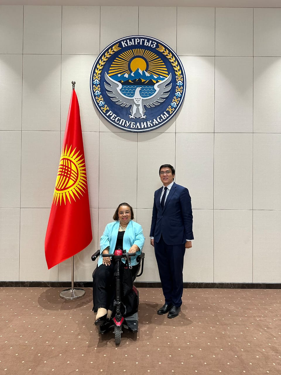 This image shows Dr. Heba Hagras and the Deputy Prime Minister of Kyrgyzstan standing in front of the official government seal and flag, in the presidential building of Kyrgyzstan. The seal on the wall is the national emblem of Kyrgyzstan. It features a golden eagle with spread wings, mountains, a sun, and text in the Kyrgyz language. To the left of the seal is the national flag of Kyrgyzstan, which is red with a yellow sun design in the center. In the foreground are two people: 1. Dr. Heba Hagras in a wheelchair, wearing a light blue jacket and black pants. 2. The Deputy Prime Minister standing next to her, wearing a dark suit. They are posing for an official photo. The image conveys a sense of formal diplomacy or official government business, related to international relations and disability rights.