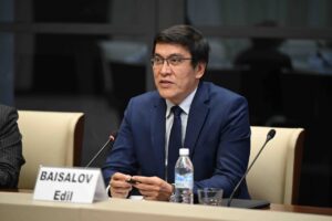 image shows a man in a dark blue suit speaking at a similar type of event. There's a nameplate visible that says "BAISALOV Edil".