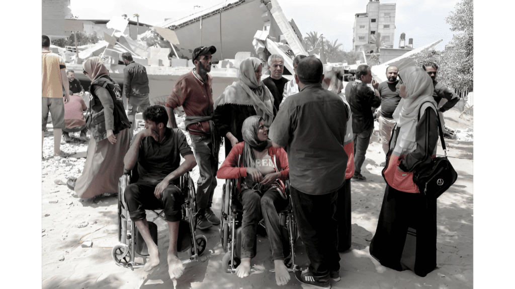 This image is from a conflict or disaster zone in Palestine, showing civilians in the aftermath of a building being destroyed. In the foreground, there are two people in wheelchairs, with several other people standing around them. Many of the women are wearing headscarves, and there appears to be rubble and destroyed buildings in the background. The people appear to have been displaced or affected by whatever event caused the destruction. The image conveys the human impact of conflict or disaster on vulnerable populations, including those with disabilities.