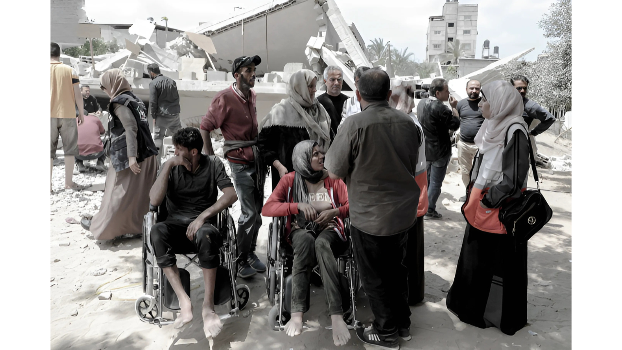This image is from a conflict or disaster zone in Palestine, showing civilians in the aftermath of a building being destroyed. In the foreground, there are two people in wheelchairs, with several other people standing around them. Many of the women are wearing headscarves, and there appears to be rubble and destroyed buildings in the background. The people appear to have been displaced or affected by whatever event caused the destruction. The image conveys the human impact of conflict or disaster on vulnerable populations, including those with disabilities.