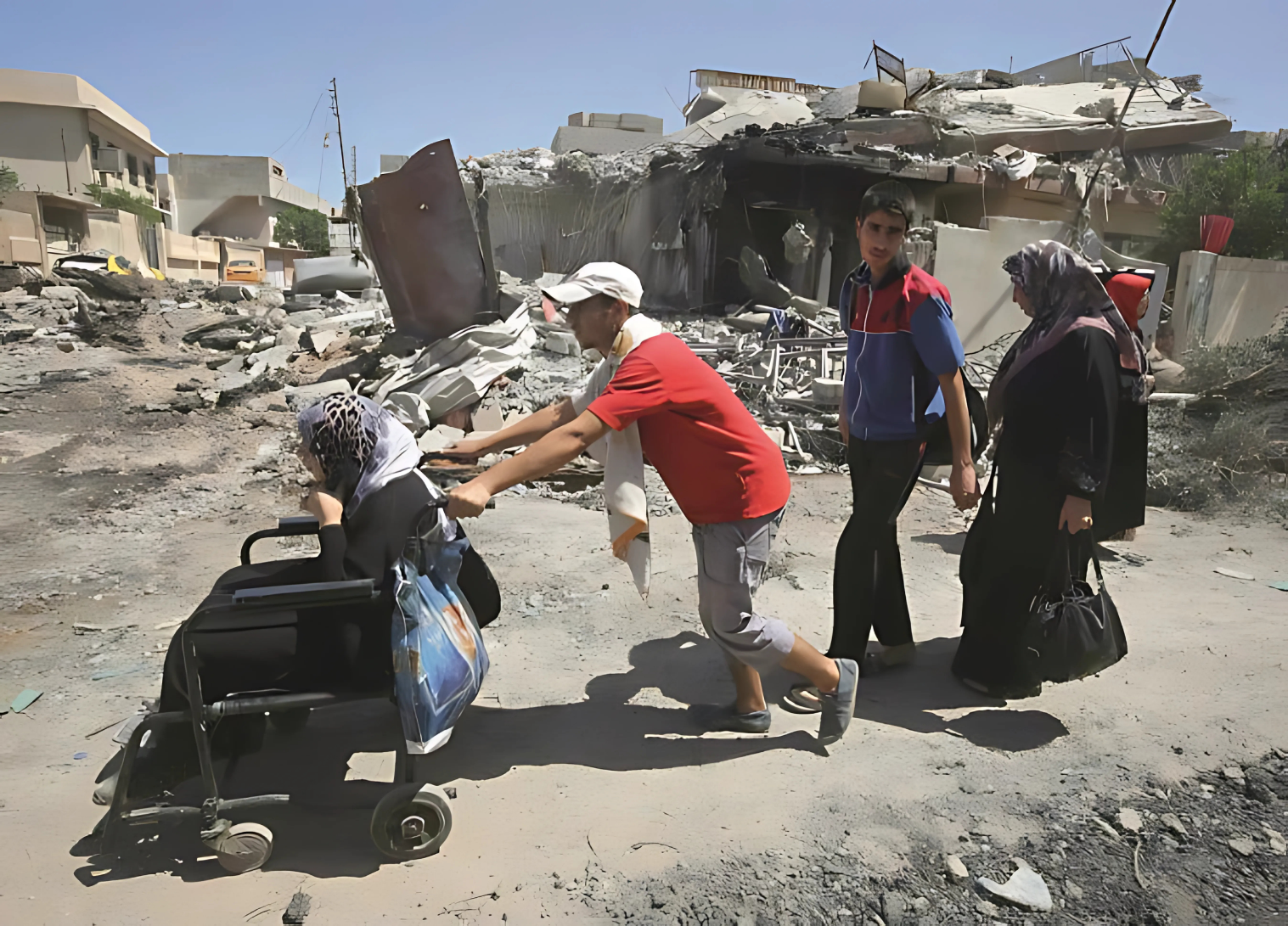 The image depicts a scene of destruction and displacement, in Palestine. In the foreground, there are several people, including: 1. A woman in a wheelchair being pushed by another person wearing a red shirt and white hat. 2. A young man wearing a blue and red shirt standing nearby. 3. A woman wearing a black hijab standing to the right. Behind them, there is extensive damage to buildings and infrastructure. A large building appears to have collapsed or been destroyed, with rubble and debris scattered around it. The surrounding area shows signs of destruction, with partially standing walls and damaged buildings visible. The scene suggests that these individuals are civilians fleeing or moving through an area that has seen significant destruction, possibly due to conflict. The presence of the wheelchair suggests that they may be assisting vulnerable individuals to evacuate or move through the affected area. The image conveys a sense of hardship and the human impact of destruction, whether from war or other causes. The report highlights the challenges faced by civilians in such situations, particularly those with limited mobility.