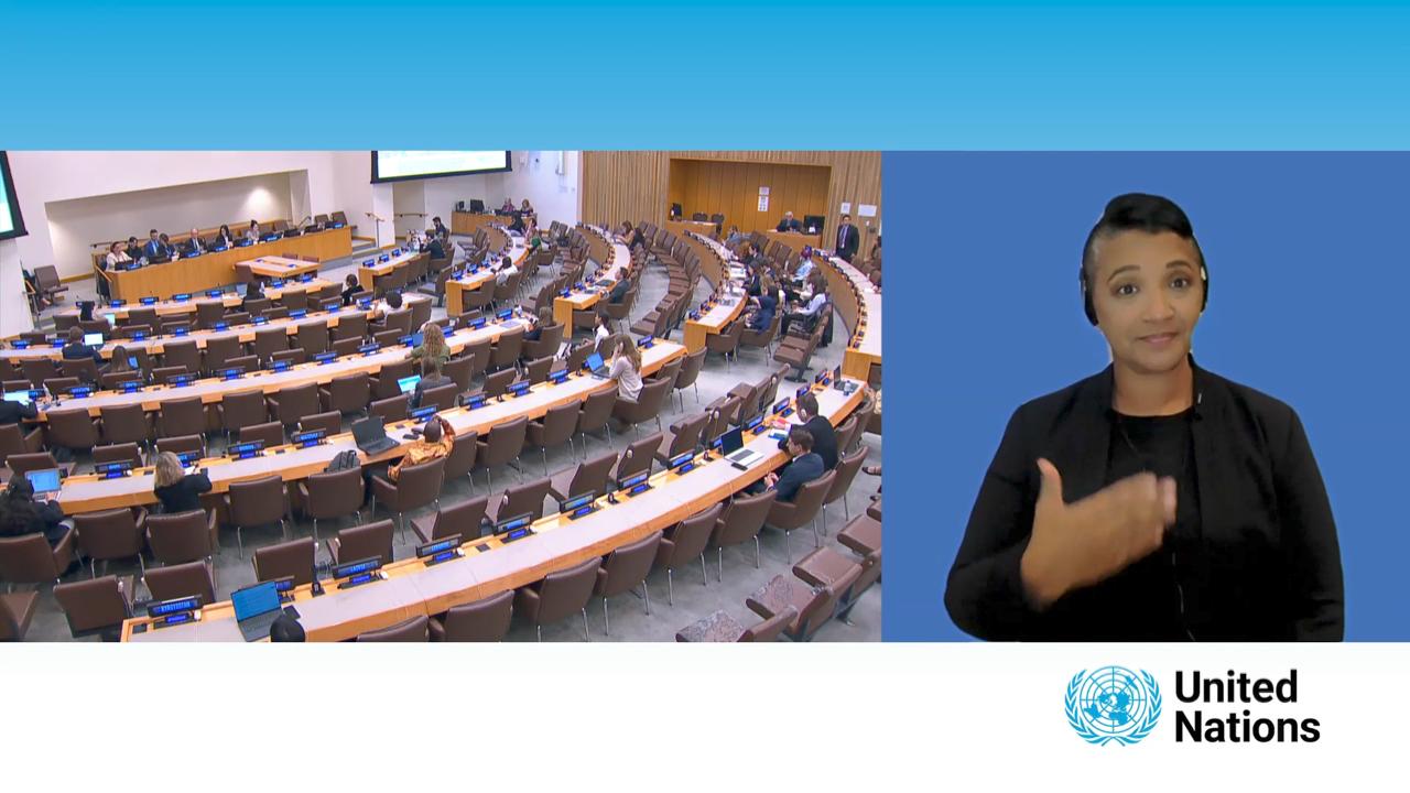 The image depicts a United Nations conference room with several participants seated at curved rows of desks equipped with microphones and nameplates. There is a screen at the front, and people are scattered around the room, suggesting an active yet orderly session. On the right side of the image, there is an interpreter providing sign language interpretation, and the United Nations logo is displayed at the bottom of the image. The setting indicates an official UN meeting, emphasizing inclusivity with sign language interpretation.