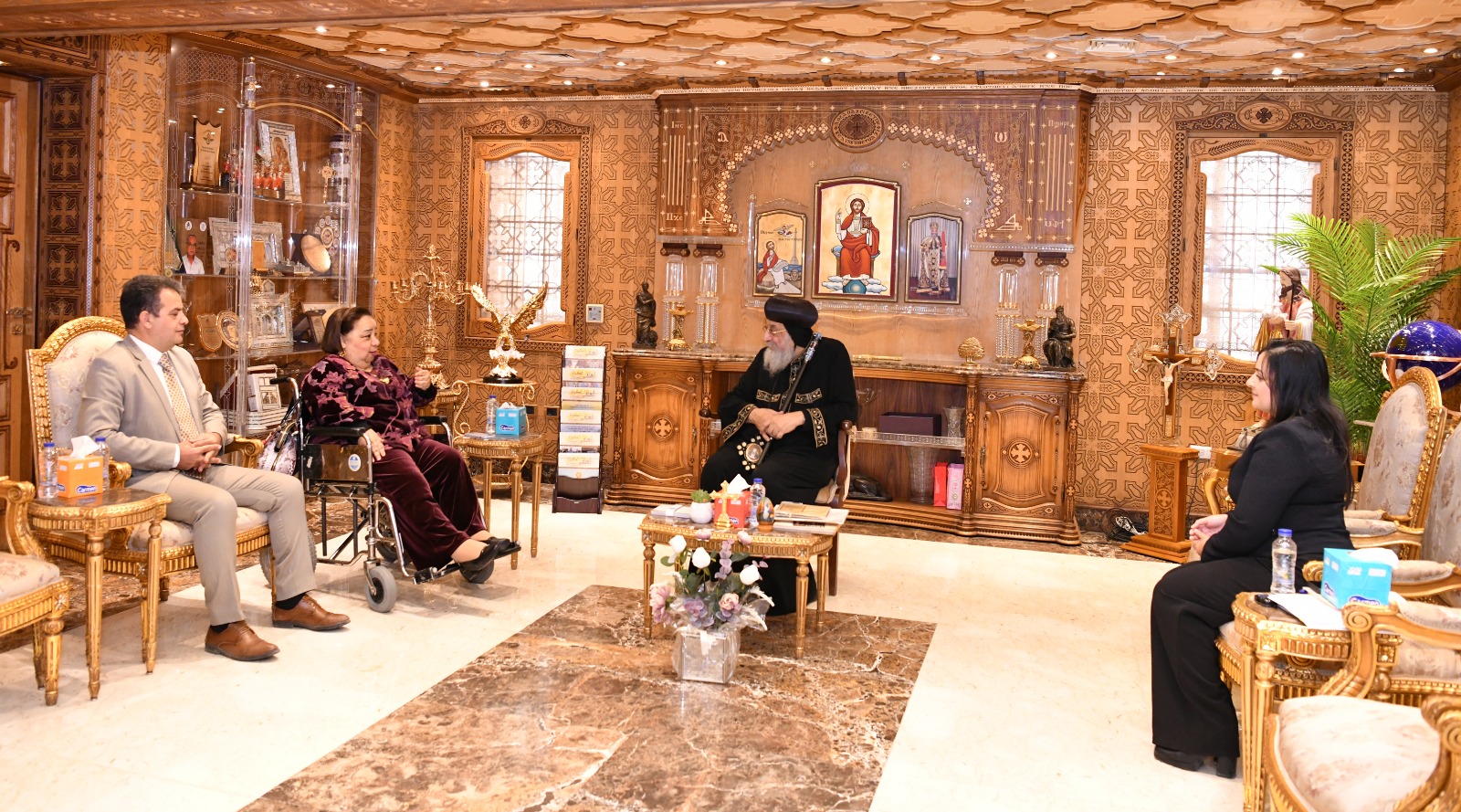This image shows a formal meeting in a room decorated with wooden ornaments and religious icons. Four people are seated, engaged in discussion. In the middle, His Holiness Pope Tawadros II is a bearded man wearing black religious garb and a head covering. To his left, the UN Special Rapporteur on the Rights of Persons with Disabilities speaks in a wheelchair wearing velvet, accompanied by a man in a beige suit. To his right, another woman dressed in black listens attentively. The room features gilded furniture, a glass display case with religious objects, and a central table with flowers and refreshments. The setting suggests a religious meeting.