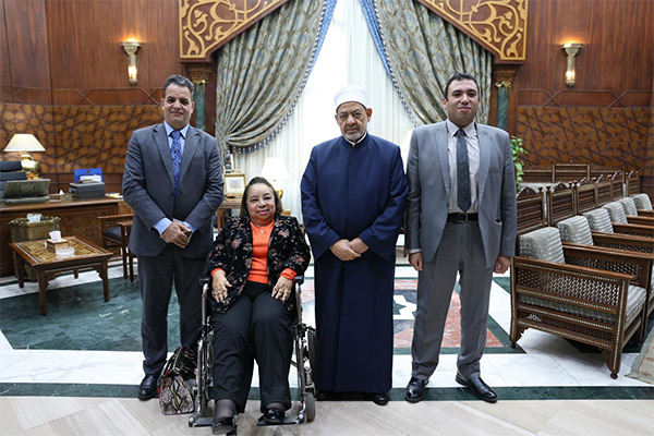 A group of four individuals are gathered in an ornate office. The group consists of the UN Special Rapporteur on Disability, seated in a wheelchair and wearing a black and orange outfit; the Grand Imam Dr. Ahmed El-Tayeb, Sheikh of Al-Azhar, dressed in traditional Islamic attire; and two men wearing suits. The background features elegant wooden panels, a large desk, and intricately embroidered curtains.